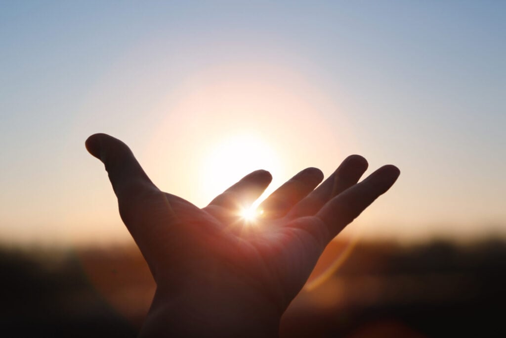 A hand is held up toward a bright rising sun, with sunlight shining between the fingers and a soft, blurred background, gently capturing a quiet moment of grief and reflection.